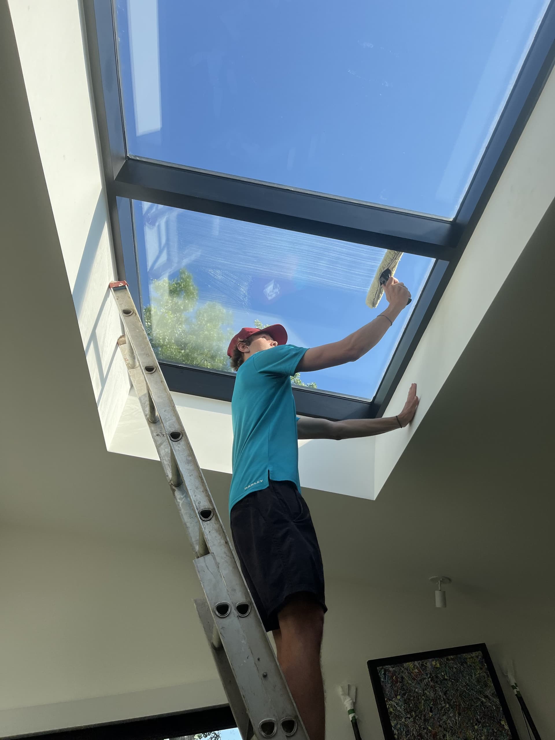 Person on a ladder cleaning a skylight with a squeegee under a clear blue sky
